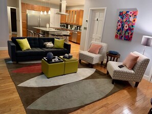 A modern living room with a sofa, ottoman, and armchair, a kitchen in the background, and a colorful painting on the wall. - MODERN Spacious 4bed 2bath HARVARD MIT BOS Cambridge Somerville walk everywhere! (Somerville)