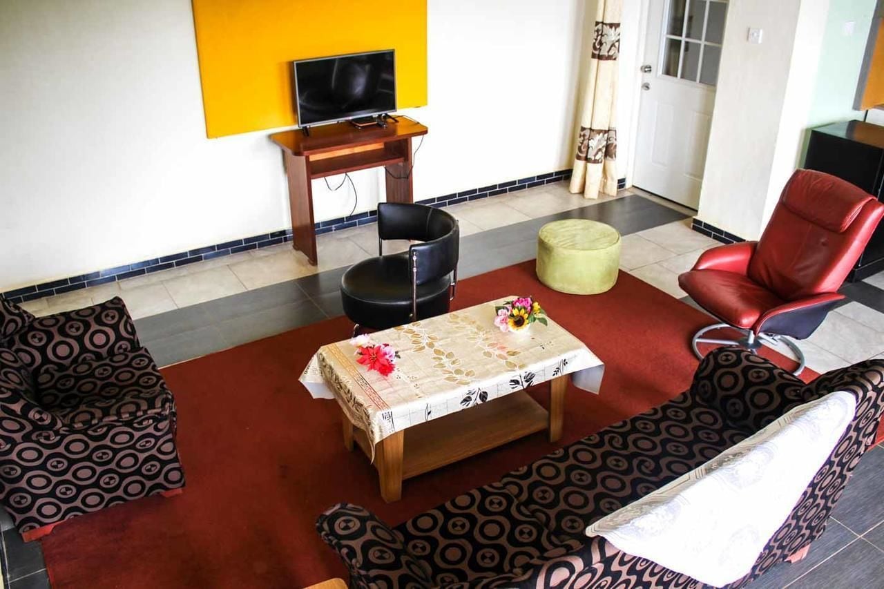 Un salon avec des canapés à motifs, une table basse ornée d’un chemin de table à fleurs, un téléviseur posé sur un meuble en bois et un fauteuil rouge.
