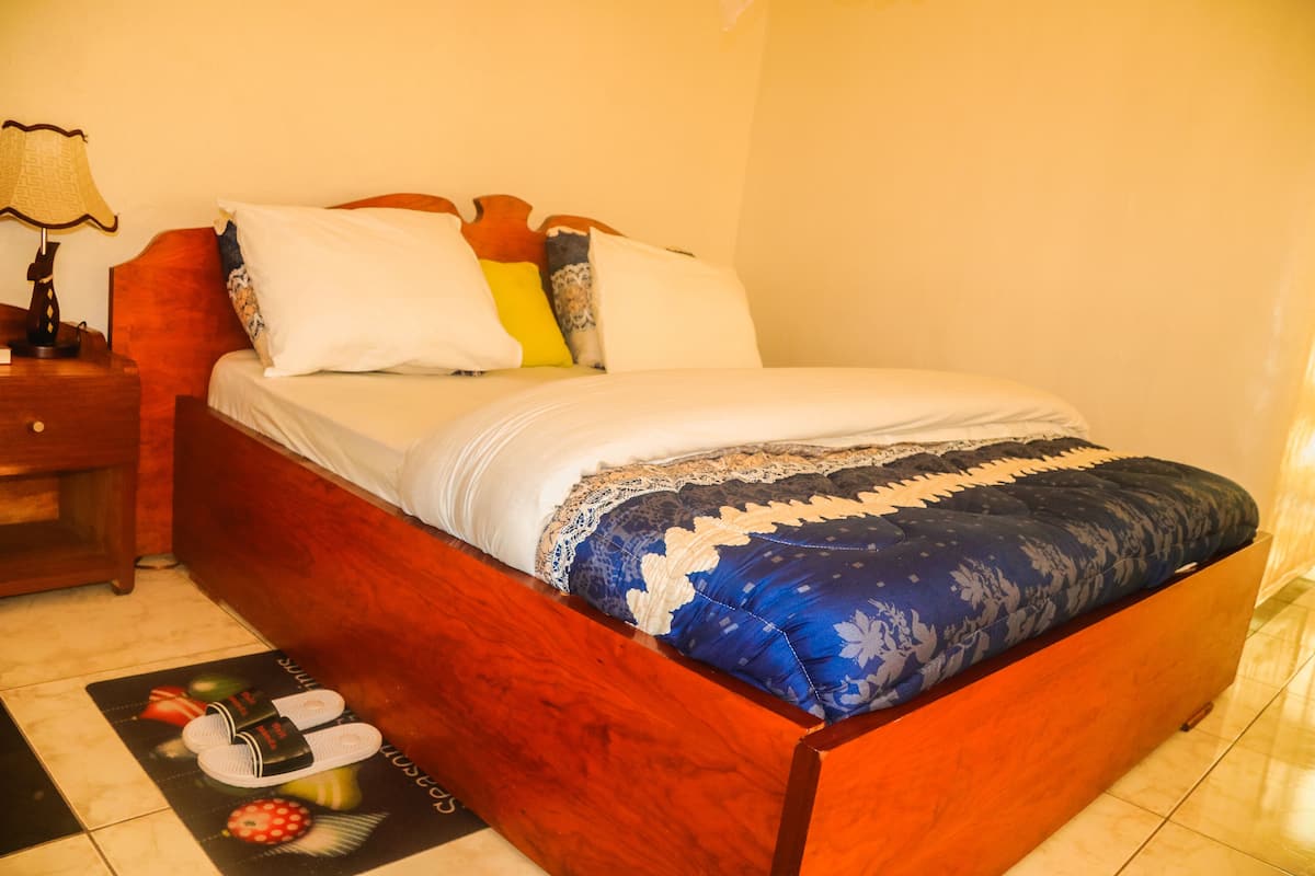 Una cama bien hecha con cabecera de madera, una mesita de noche con lámpara y un tapete con motivos de frutas.