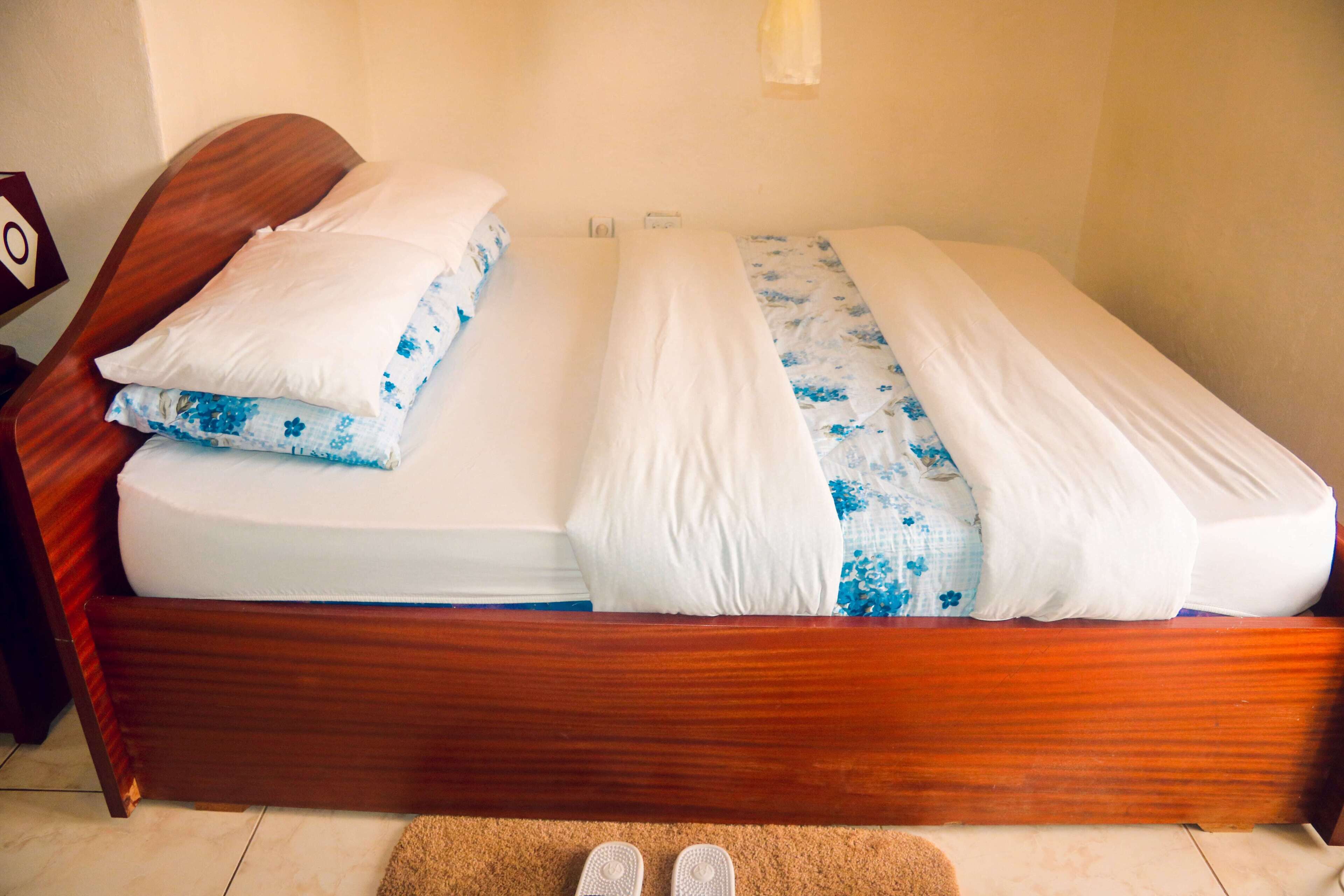 A neatly made bed with a wooden frame, white and blue floral bedding, and a pair of shoes on a small rug.