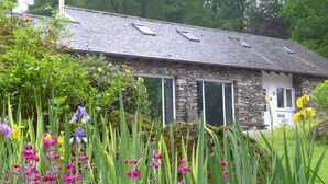 A stone house with a tiled roof, surrounded by lush greenery and flowering plants.