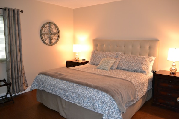 A bedroom with a bed, two bedside tables, a lamp, a wall decoration, and a window with curtains.