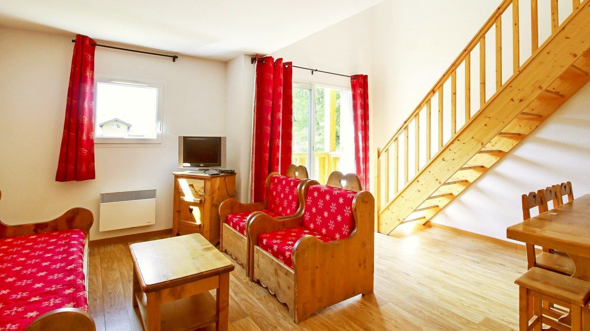 Ein gemütliches Zimmer mit einer Holztreppe, einem Fernseher und einem Essbereich mit roten Vorhängen.