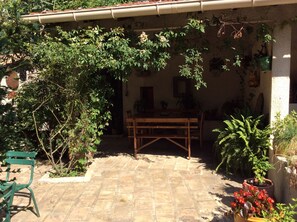 Un espace patio avec une table et des chaises en bois, entouré de plantes en pot et doté d’un sol en briques.