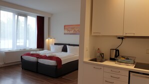 A hotel room with a bed, a window with curtains, a wall-mounted lamp, a painting, a sink with a kettle, and a countertop with a kettle and a dish rack. - Apart- Hotel Kick (Sinsheim)