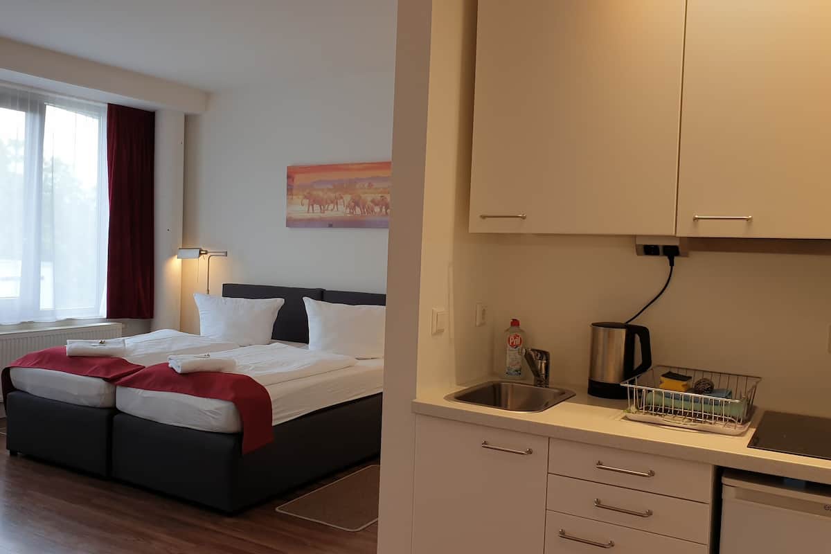 Une chambre d’hôtel avec un lit, une fenêtre avec des rideaux, une lampe fixée au mur, un tableau, un lavabo avec une bouilloire, et un plan de travail avec une bouilloire et un égouttoir.