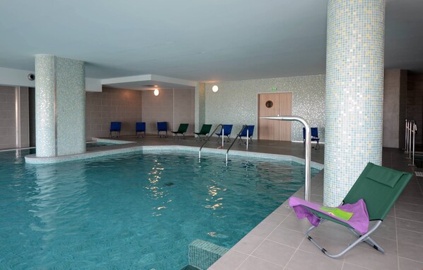 An indoor swimming pool with clear water, surrounded by lounge chairs and a tiled wall.