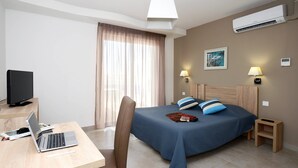A hotel room with a bed, a desk with a laptop, a television, and an air conditioning unit.