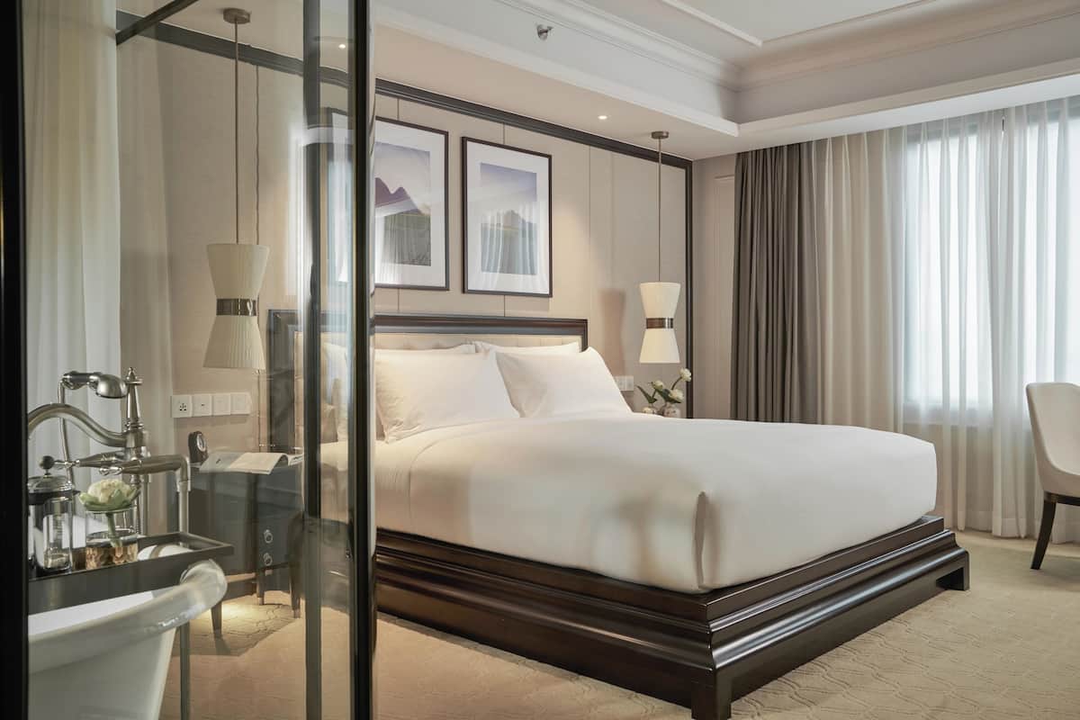 A hotel room with a large bed, a glass partition, a desk with a lamp, and a chair.