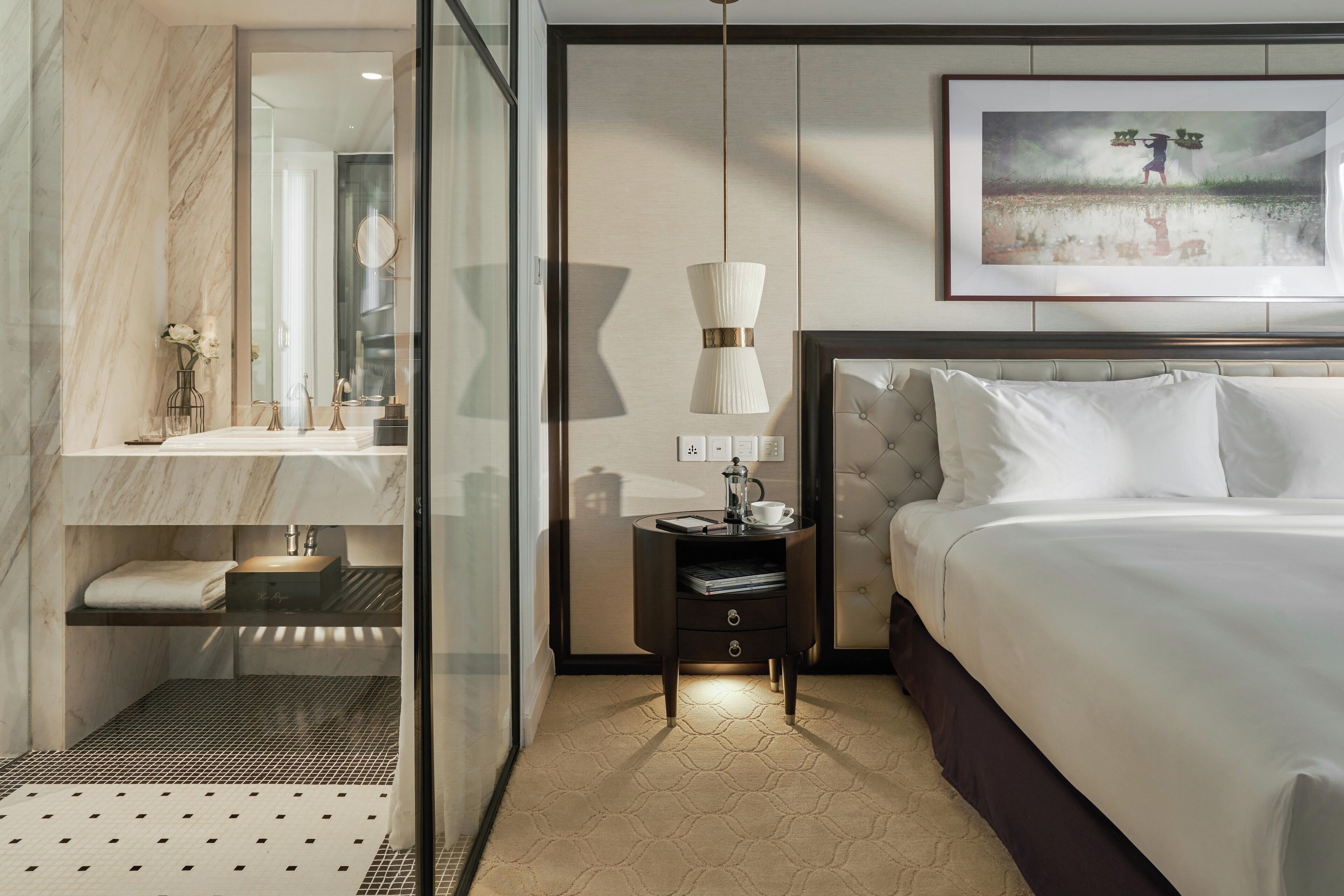 A modern hotel room with a large bed, a bedside table with a lamp, a mirror, and a framed artwork on the wall.