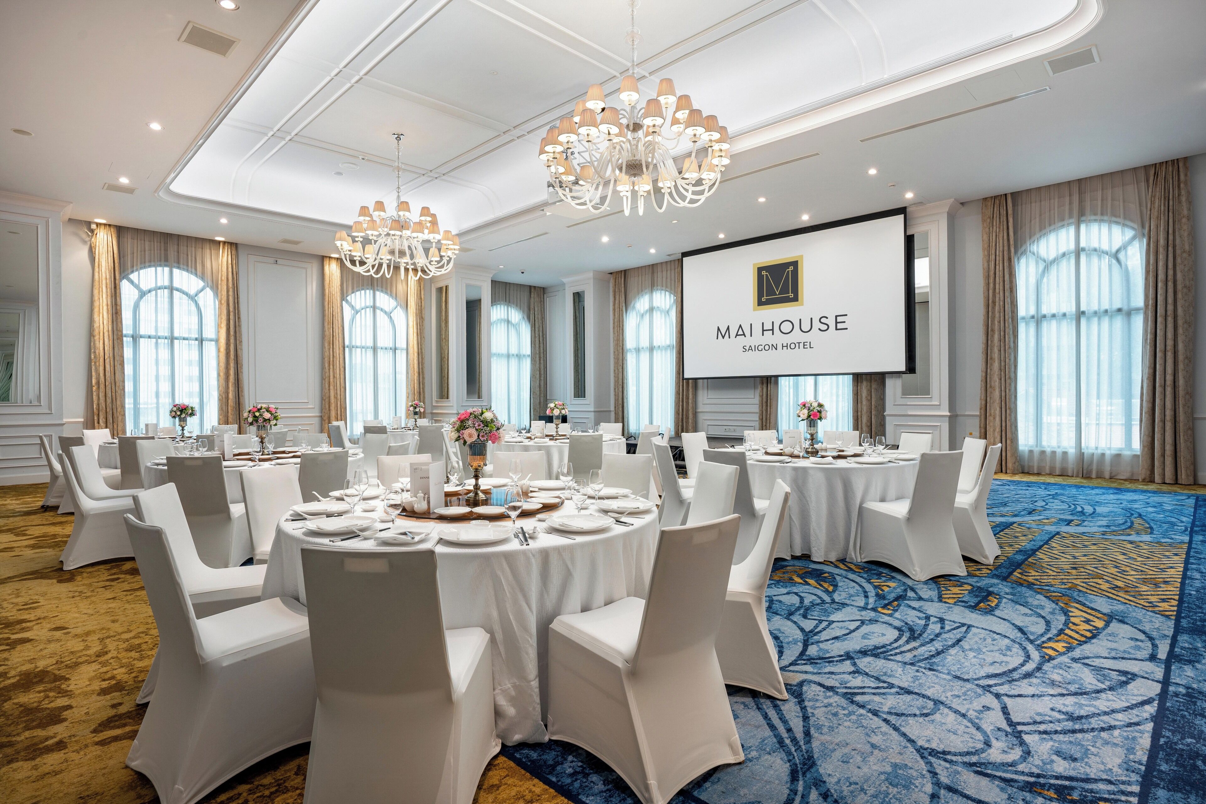 A conference room at Saigon Hotel with round tables, white chairs, chandeliers, and a large screen displaying the hotel name.
