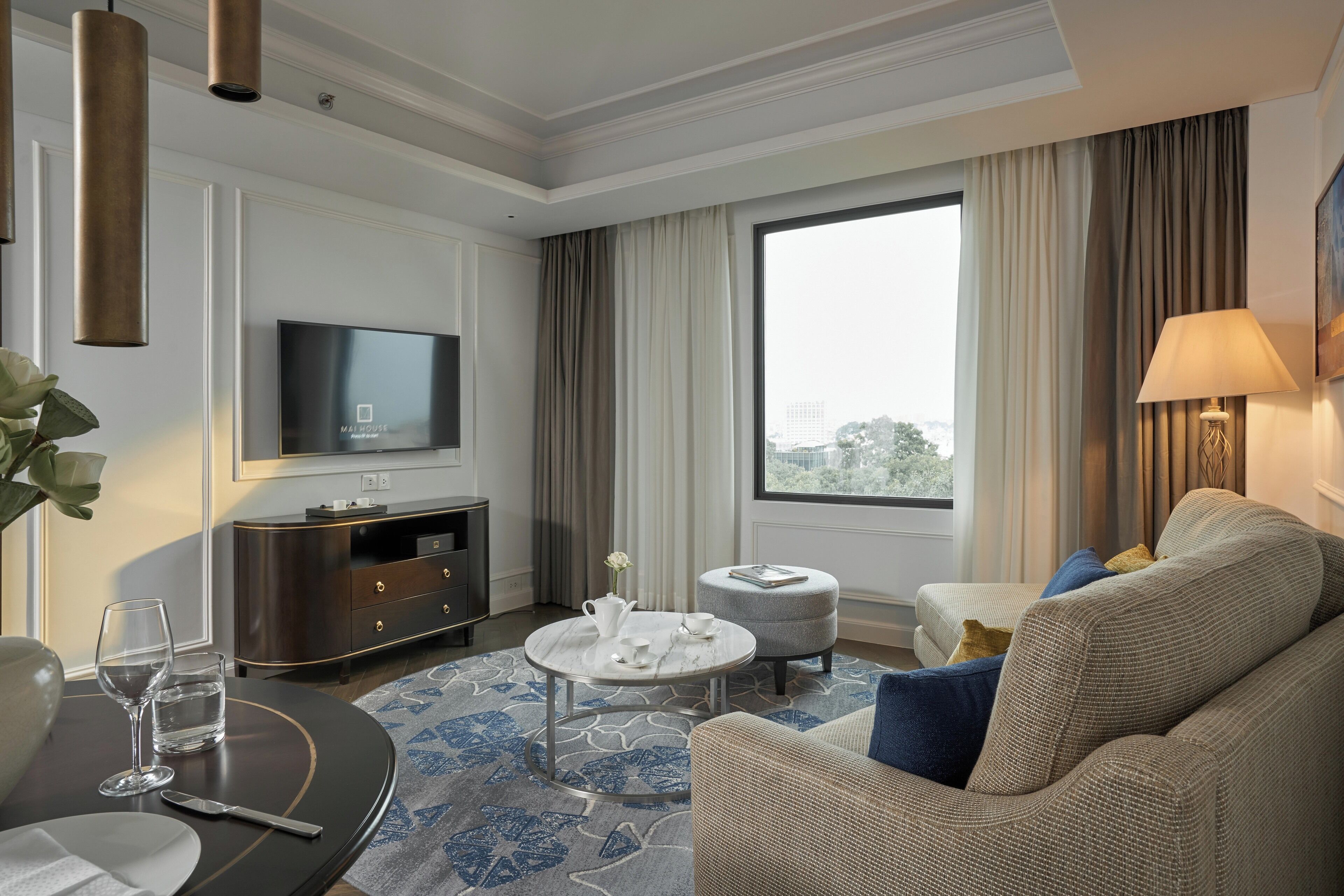 Une chambre d’hôtel moderne dotée d’une télévision à écran plat, d’une table basse ronde, d’un canapé avec des coussins, d’une table d’appoint et d’une grande fenêtre offrant une vue sur la ville.