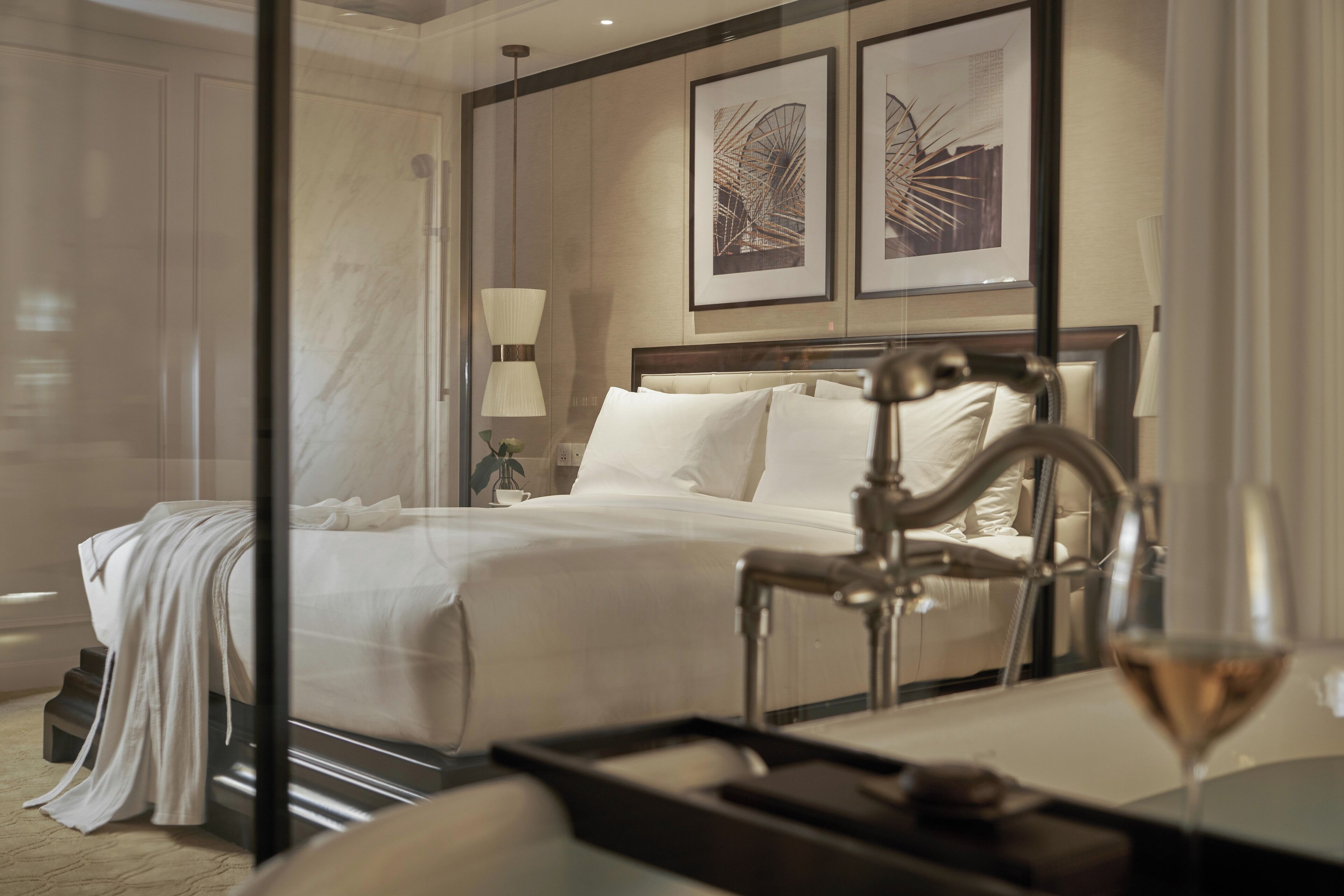 A hotel room with a bed, a bedside table with a tray, a glass of wine, and framed artwork on the wall.