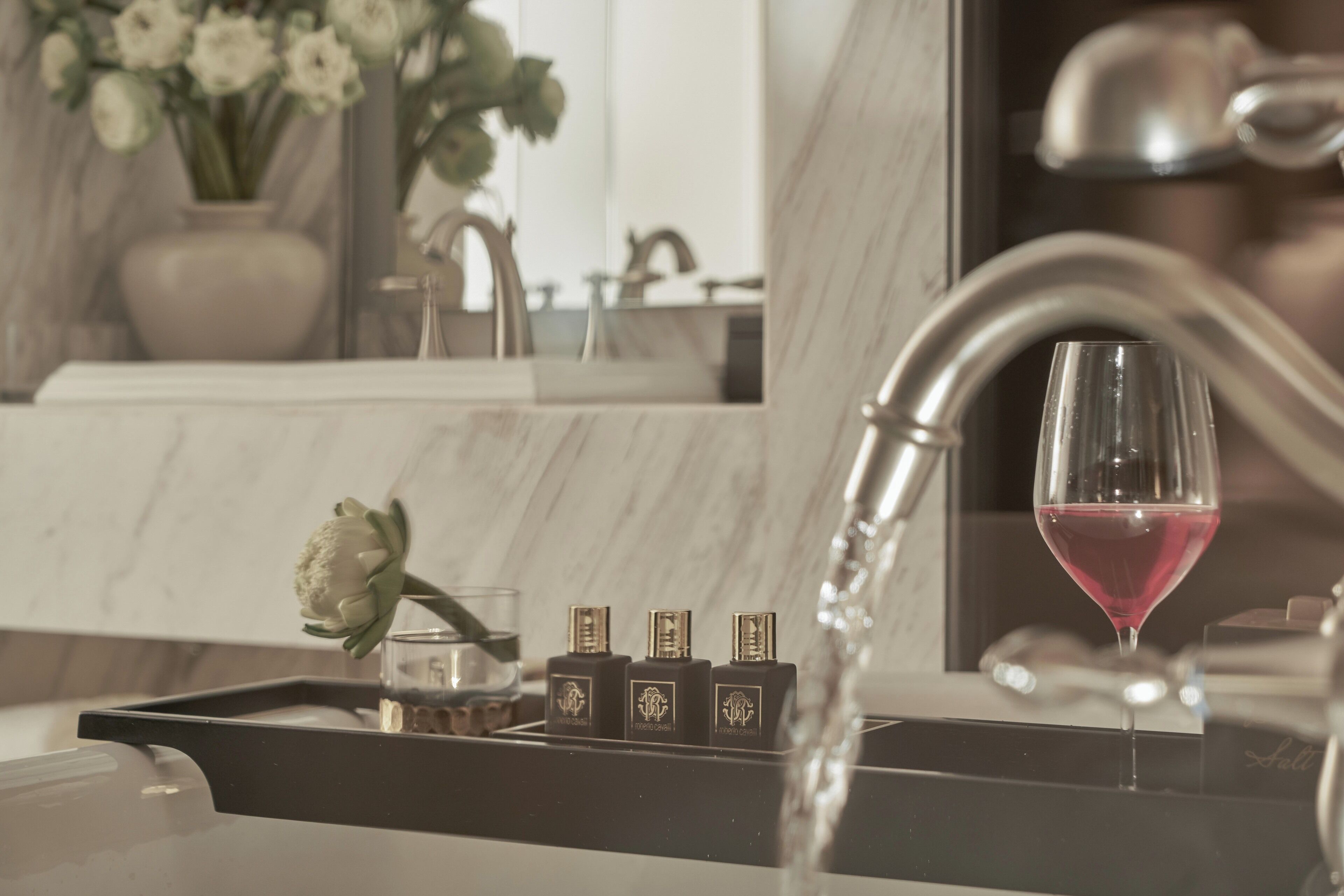 A bathroom sink with a glass of red wine, a vase with flowers, and three bottles of perfume.
