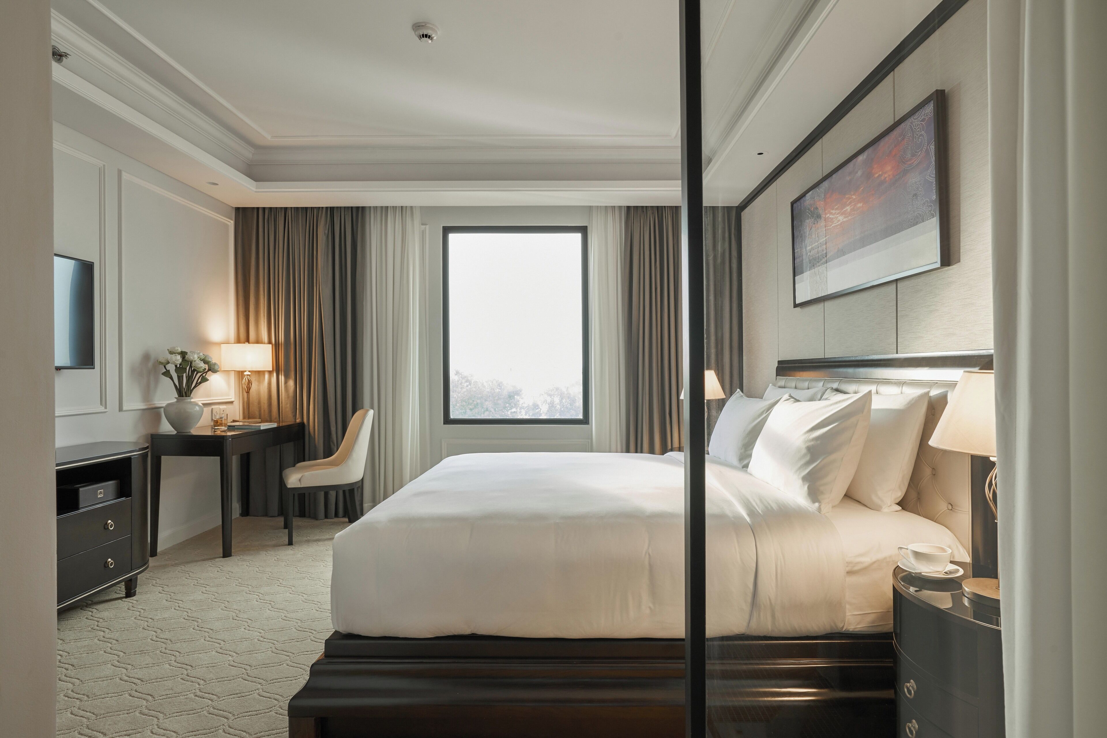 A hotel room with a large bed, a desk with a lamp, a chair, a TV, and a window with curtains.