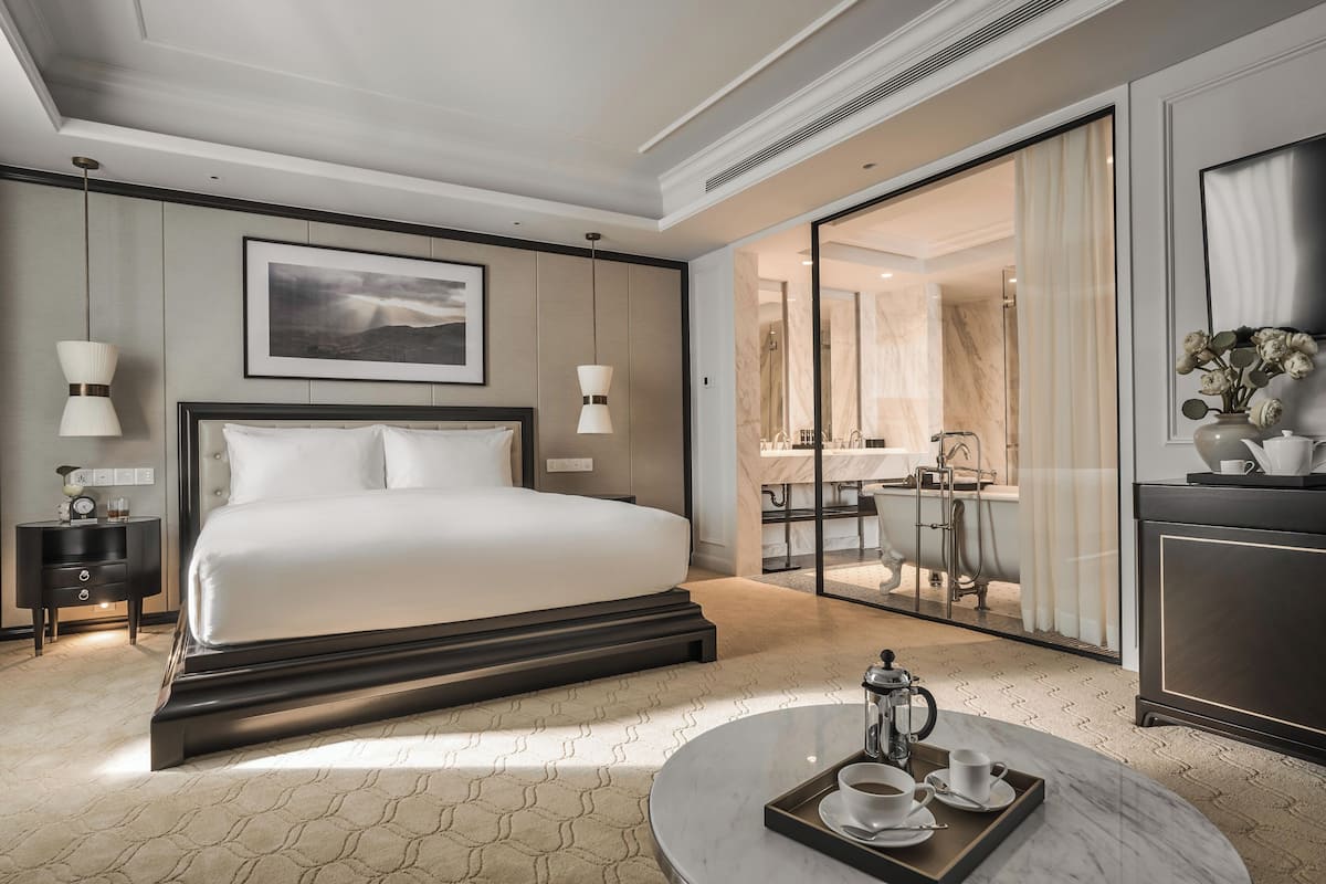 A modern hotel room with a large bed, a bedside table with a lamp, a coffee table with a tray, and a view of the bathroom through a glass door.
