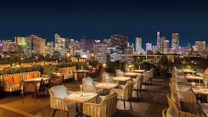 Un bar situé sur un toit, avec des meubles et des tables en osier, offrant une vue sur le panorama urbain illuminé la nuit.