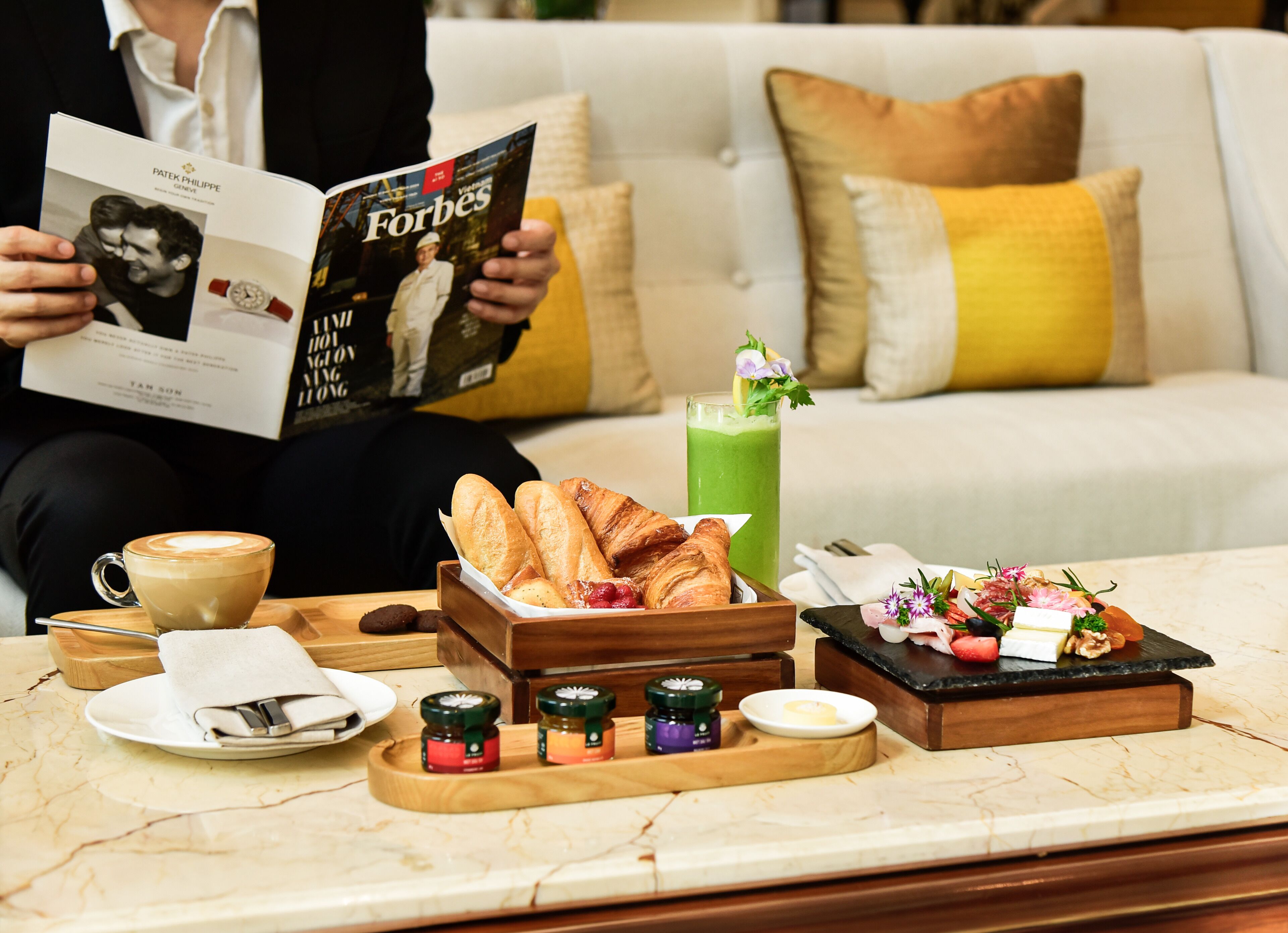 Una persona legge una rivista Forbes, una tazza di caffè, croissant, succo verde e un vassoio per la colazione con marmellate e burro.