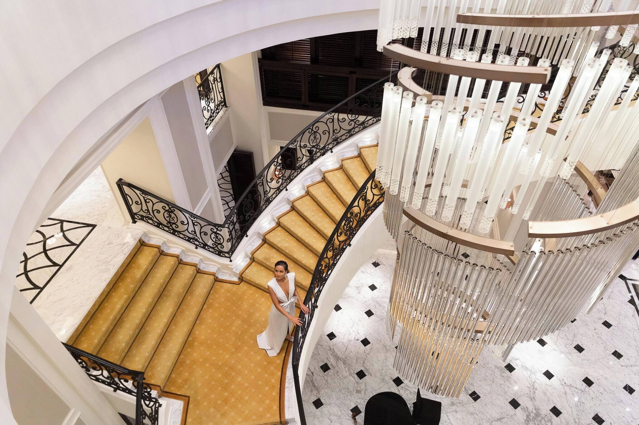 A grand staircase with a woman descending, marble floors, and a decorative chandelier.
