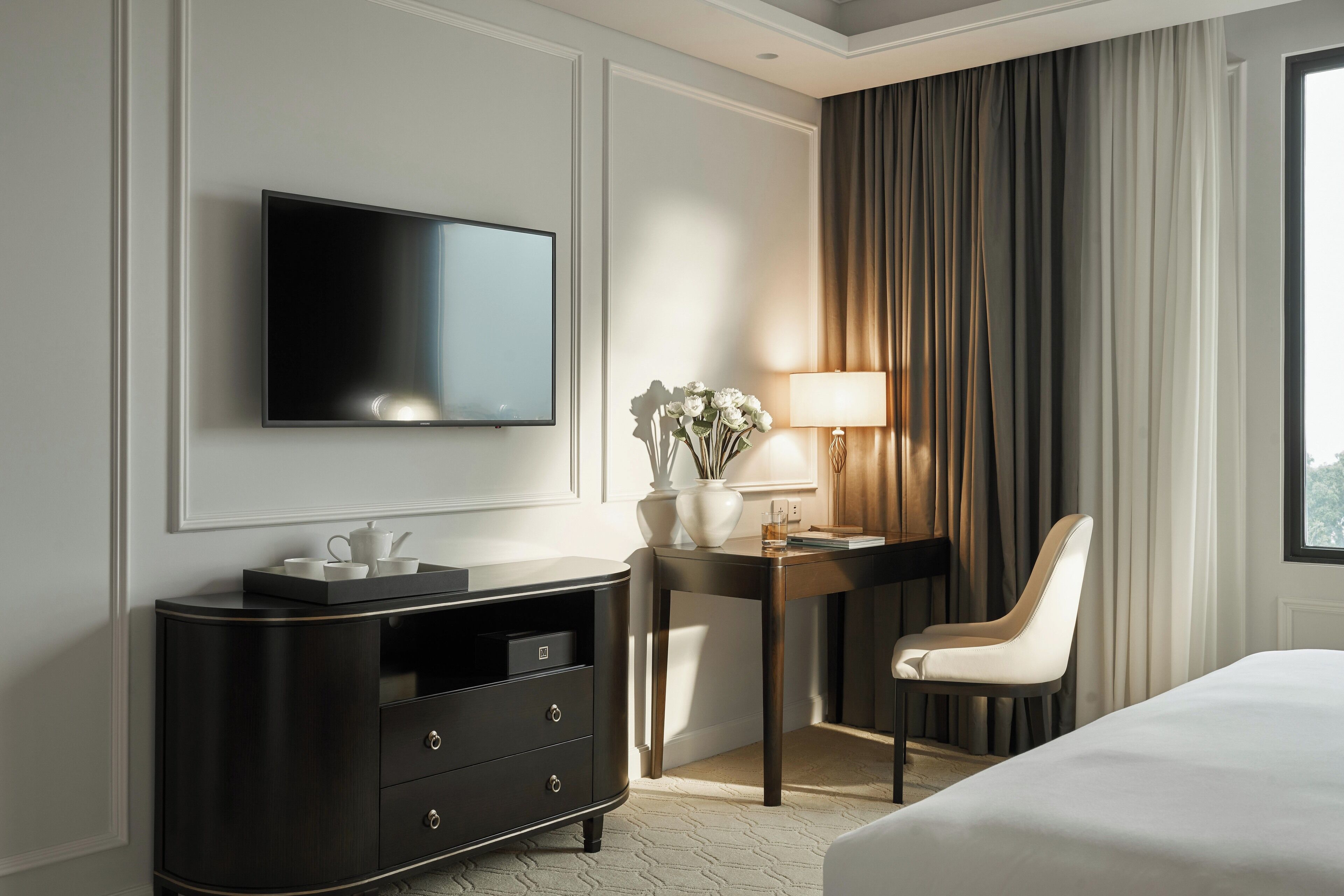 A hotel room with a flat-screen TV mounted on the wall, a dark wooden desk with a lamp, a chair, and a bed.