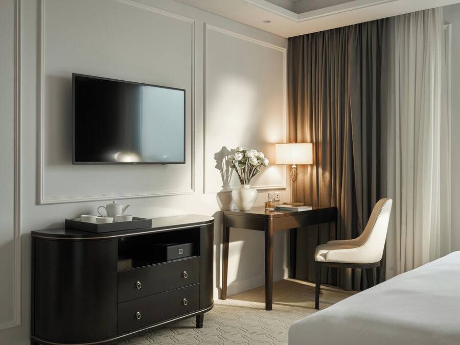 A hotel room with a flat-screen TV mounted on the wall, a dark wooden desk with a lamp, a chair, and a bed.