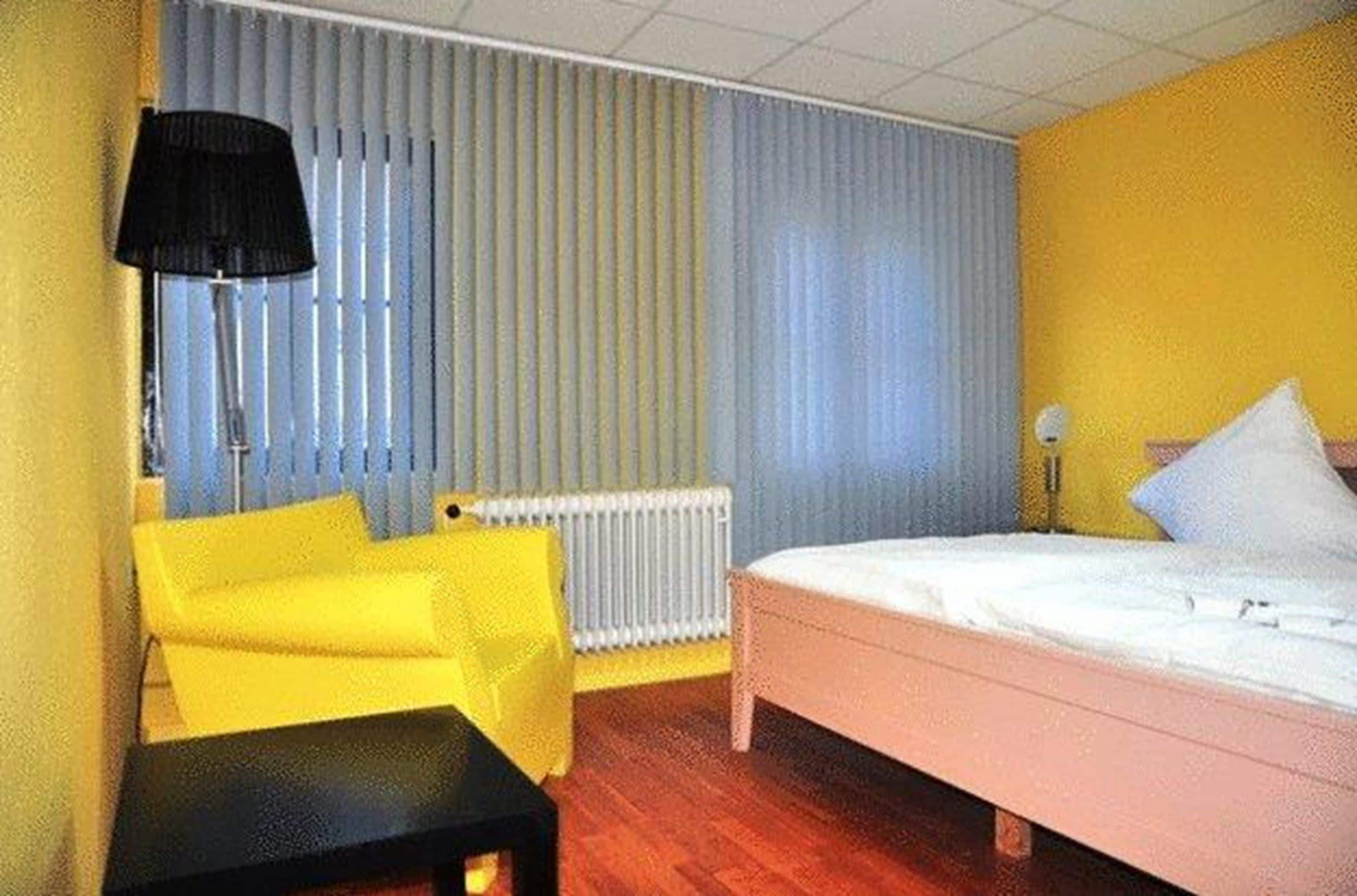 Habitació d'hotel amb un llit, un butaca groga, una taula petita, una làmpada i un radiador.