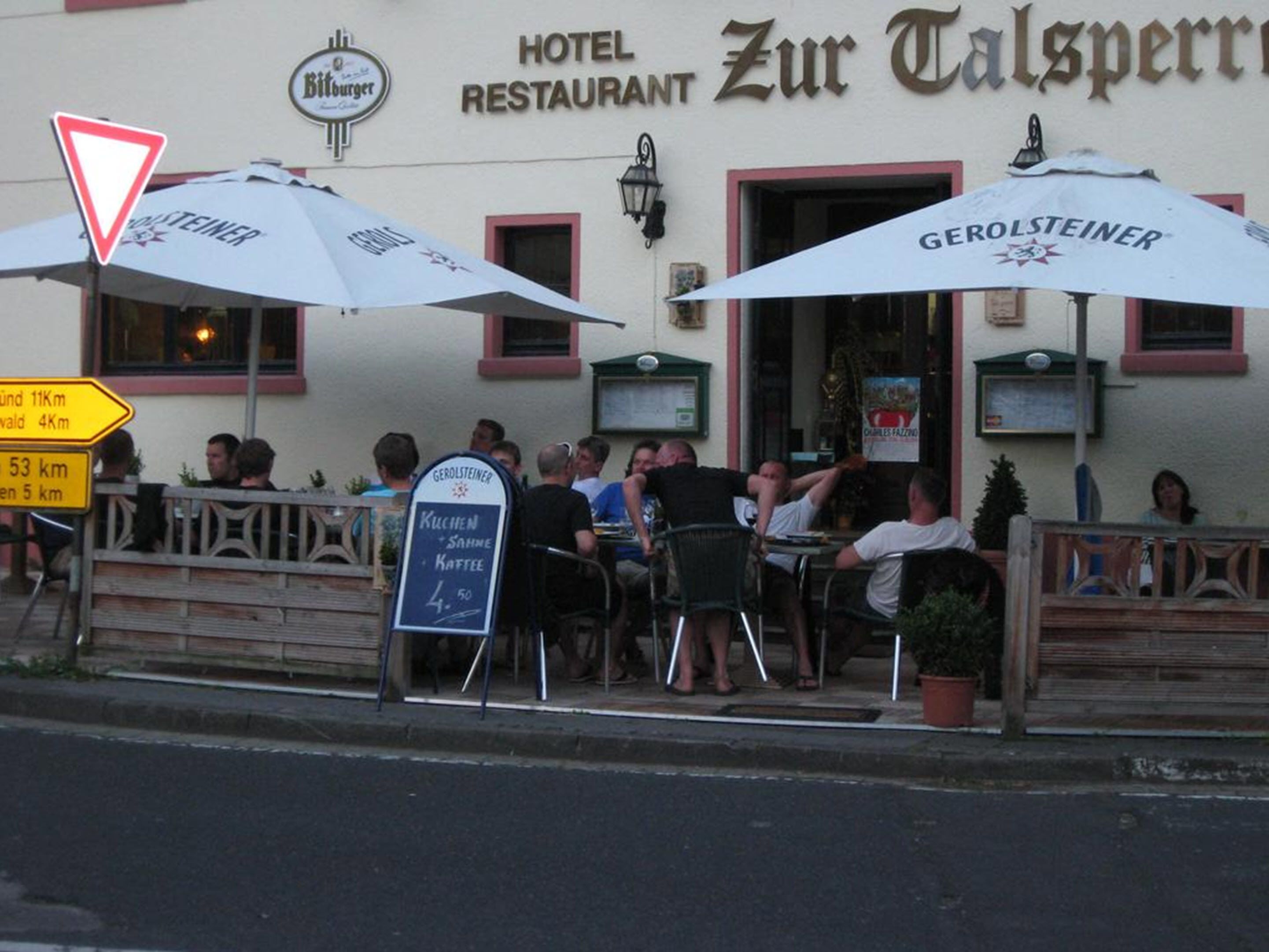 Restaurante com mesas ao ar livre, um anúncio para a cerveja Gerolsteiner e um quadro de cardápio.