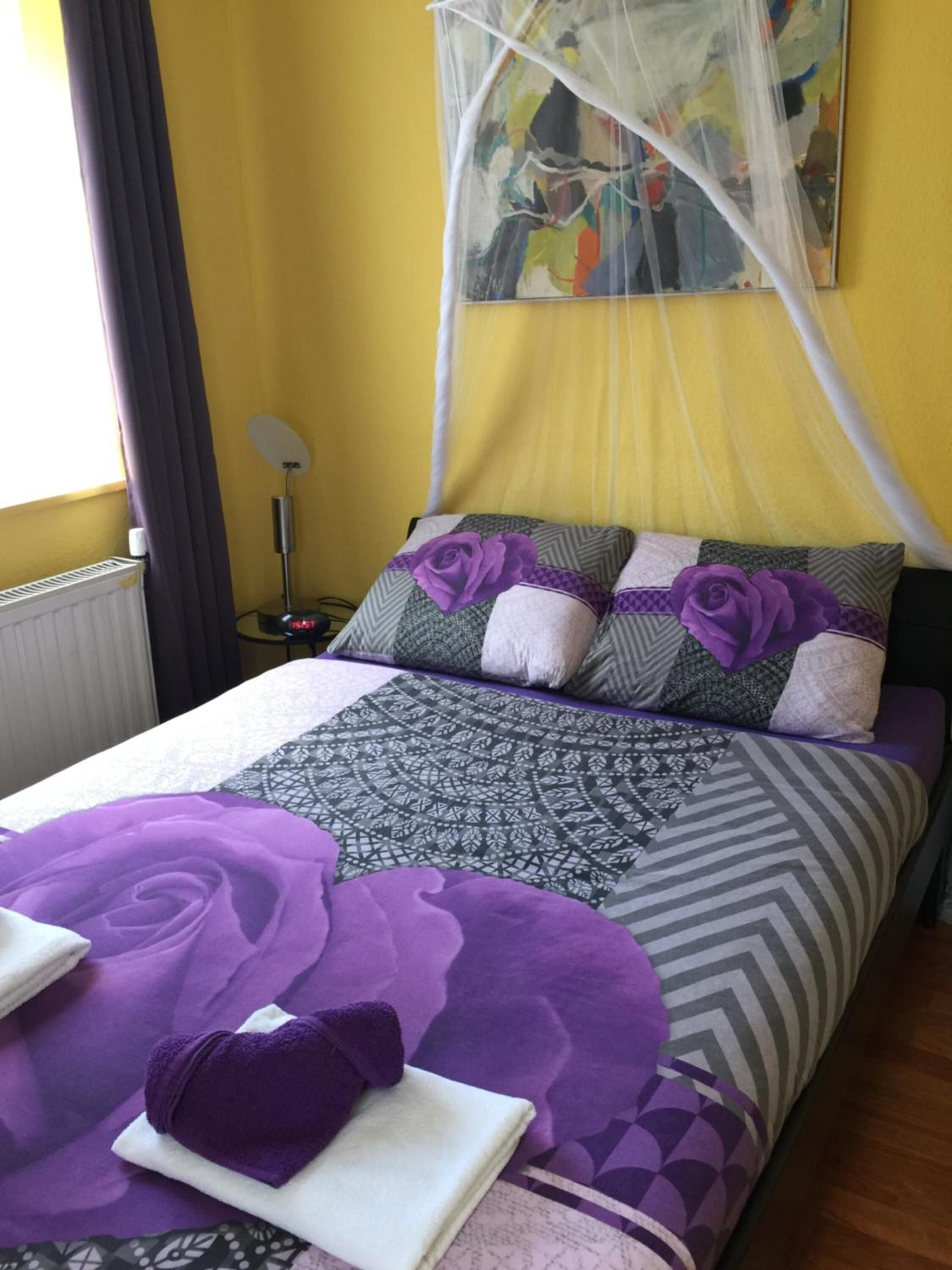 Un lit avec une couvre-lit à motifs violets et gris, une rose violette en guise de décoration et un oreiller blanc.