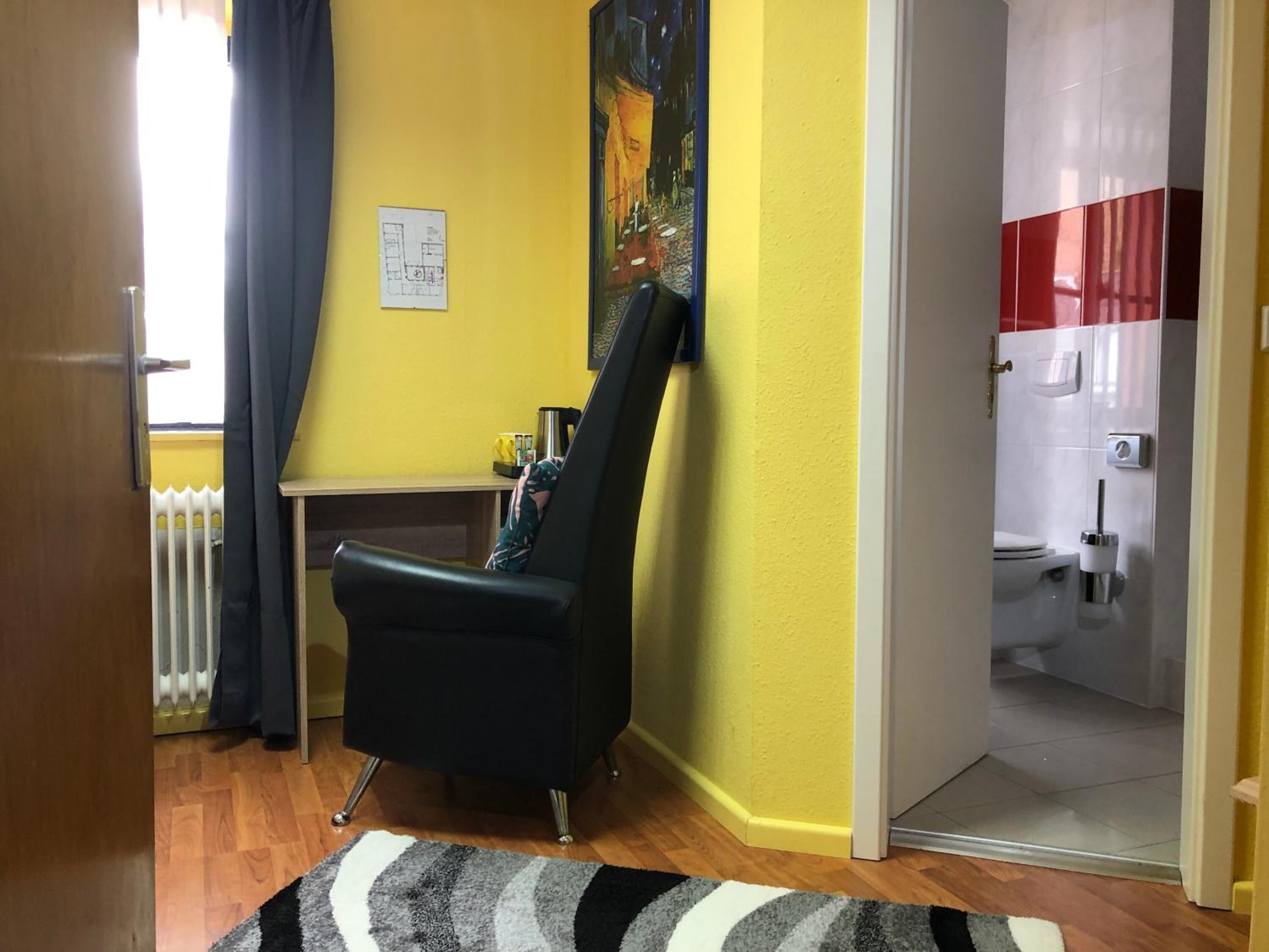 A room with a yellow wall, a black chair, a radiator, a small table with a coffee maker, and a bathroom with a toilet and sink.