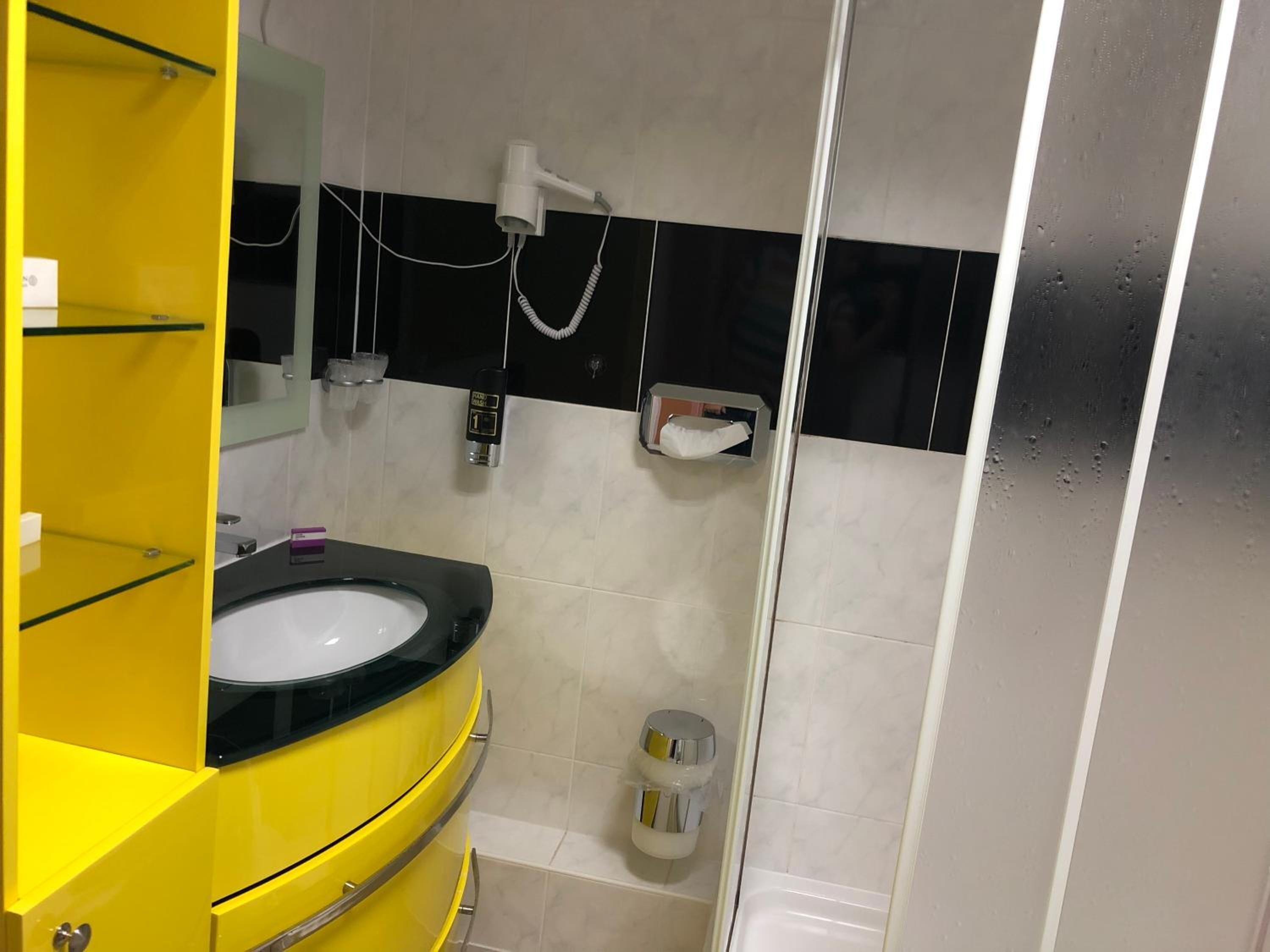 A bathroom with a yellow vanity, a black sink, a mirror, a towel dispenser, and a shower area.