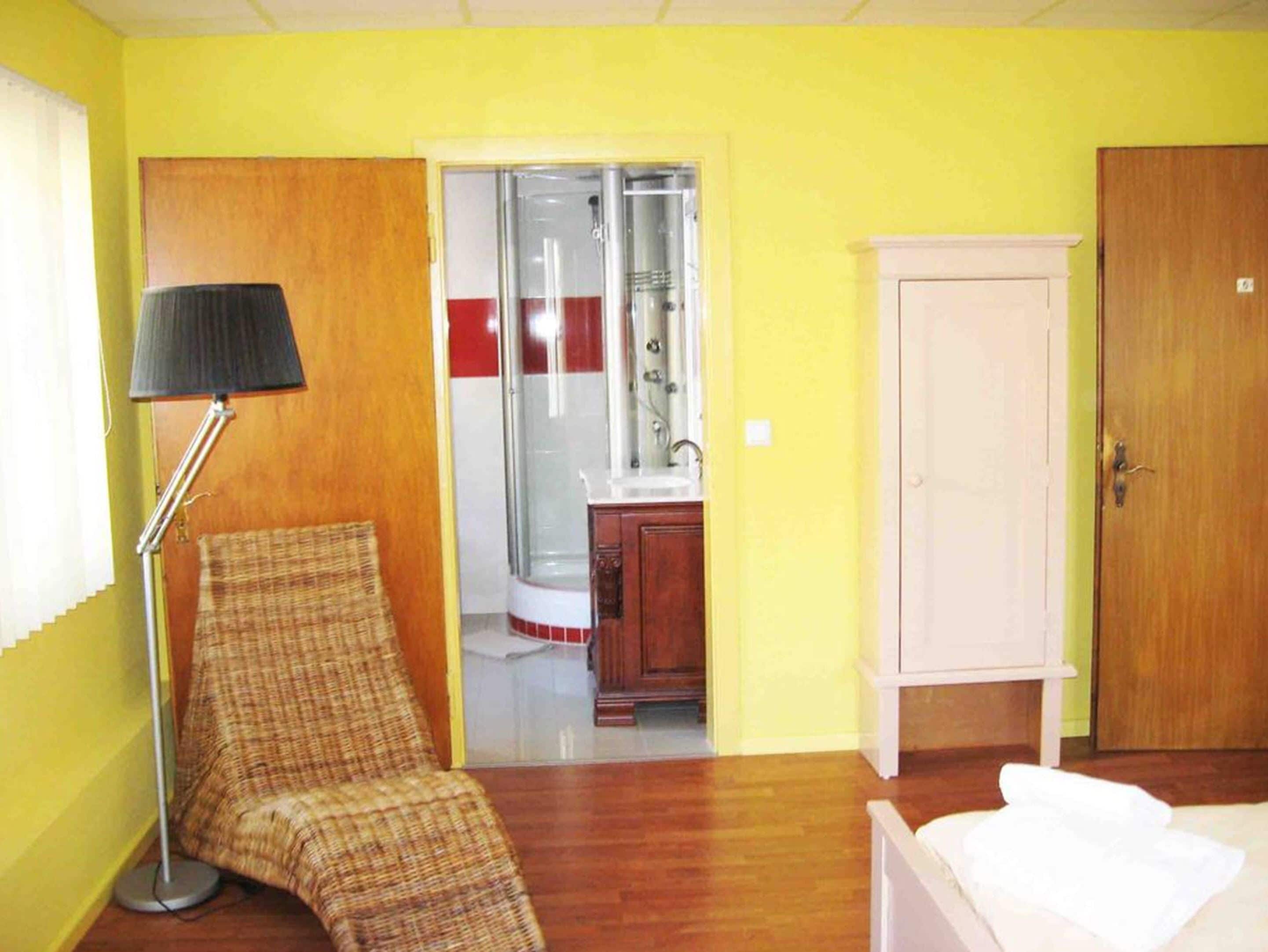 Une chambre avec un lit simple, une chaise, une lampe, une armoire et une salle de bain visible par une porte ouverte.