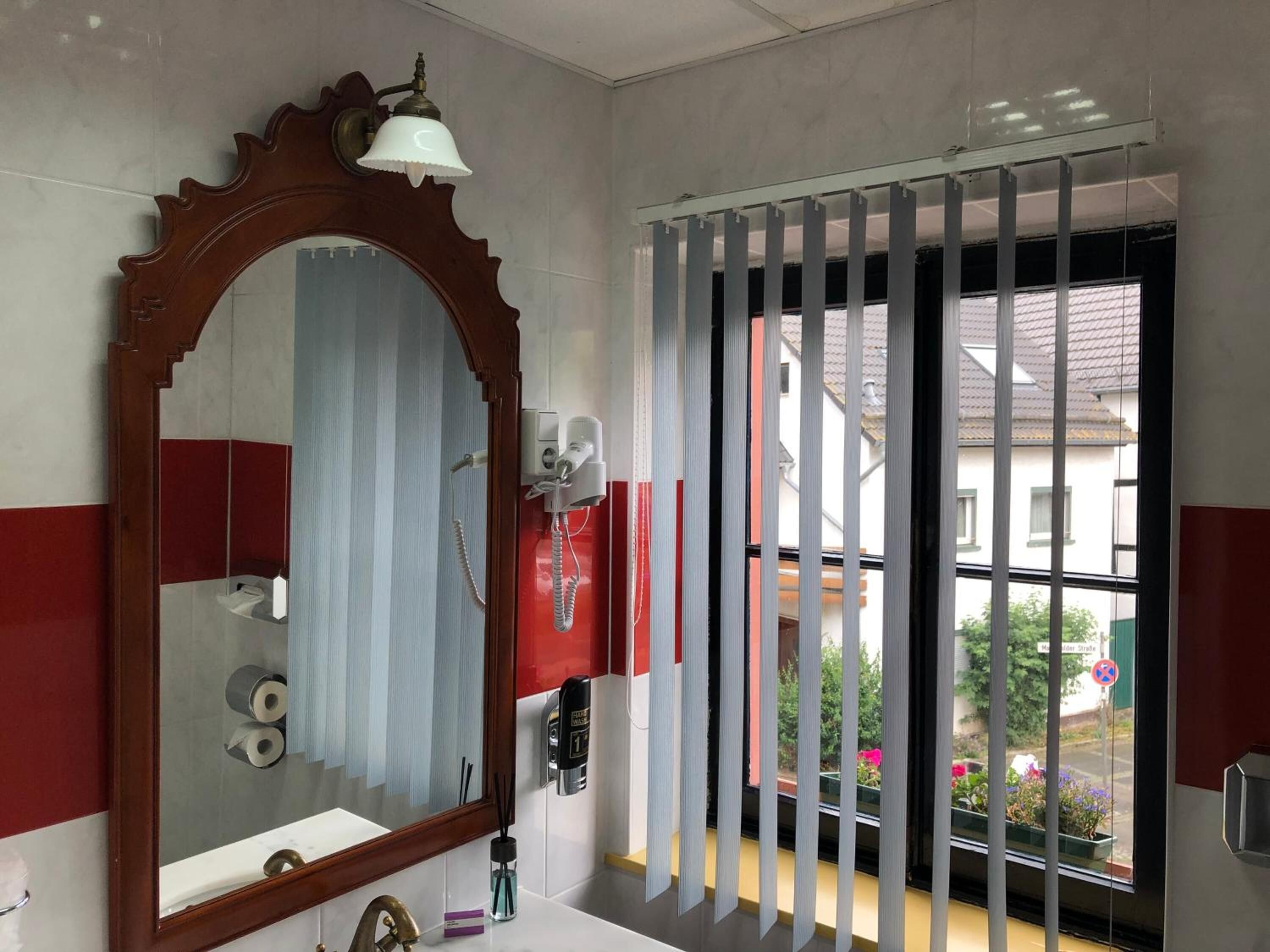 Un baño con un espejo grande, un dispensador de papel higiénico, un secador de manos y una ventana con persianas verticales.