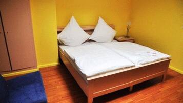 A single bed with white linens, a nightstand with a lamp, and a blue chair in a room with yellow walls and wooden flooring.