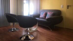 A living room with a black sofa, two black chairs, a wooden floor, and a window with vertical blinds. - Astoria Hotel Heimbach (Heimbach)