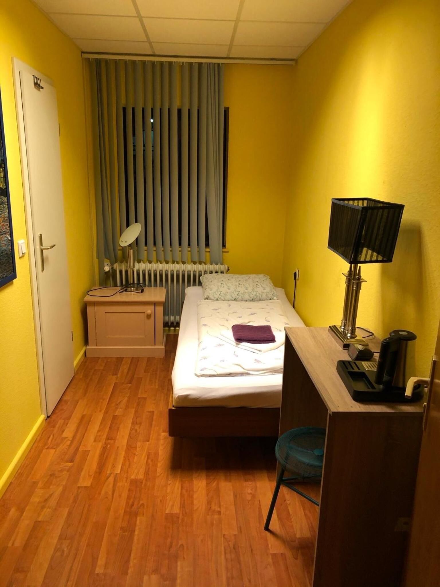 Una habitación pequeña y funcional con cama, escritorio, silla, radiador y ventana con cortinas.