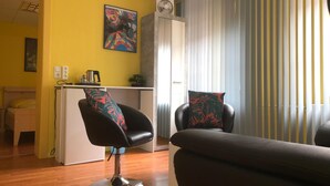 A modern living room with a yellow wall, a black leather chair with a colorful cushion, a grey sofa, and a dining area with a white counter and a painting. - Astoria Hotel Heimbach (Heimbach)