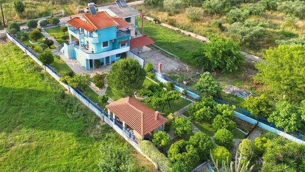 Une maison à deux étages, avec un toit recouvert de tuiles rouges, entourée d’un jardin bien entretenu et d’une clôture bleue.