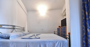 A small, well-lit bedroom with a single bed, a dresser, a television, and a curtain.