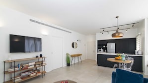 A modern living room with a flat-screen TV, bookshelves, a coffee table with fruit, a blue sofa, and a kitchen area with a dining table. - Tlv Center by Tlv2rent (Tel Aviv)