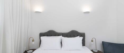 A neatly made bed with white linens, two bedside tables with lamps, a grey upholstered headboard, and a blue chair.
