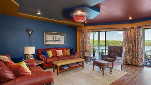 Un salón amplio con un sofá de cuero rojo, una mesa de centro de madera y un sillón a cuadros. Un gran ventanal ofrece vistas a un lago.