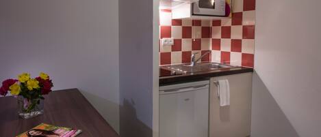 A compact kitchen with a microwave, sink, and dishwasher, a small table with a magazine and flowers, and a checkered backsplash.