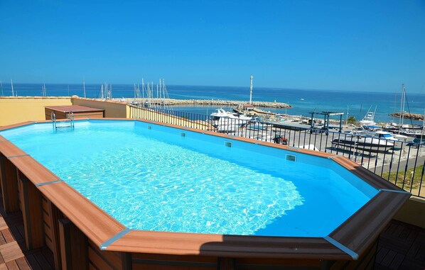 A swimming pool with a wooden deck, surrounded by a railing, overlooking a marina with boats and a clear blue sky. - Plongez dans la Nature | Appart proche de la Plage (Solenzara)