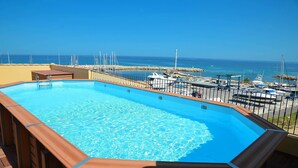 A swimming pool with a wooden deck, surrounded by a railing, overlooking a marina with boats and a clear blue sky.
