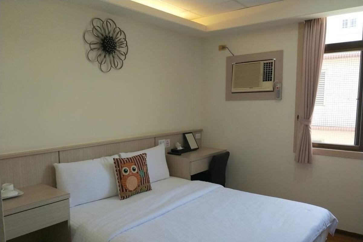 Habitación de hotel con cama, mesita de noche, escritorio, equipo de aire acondicionado y ventana con cortinas.