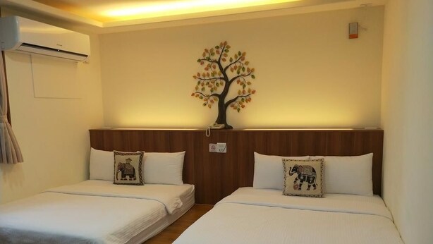 Habitación de hotel con dos camas individuales, cabecero de madera, aire acondicionado de pared y un árbol decorativo en la pared.