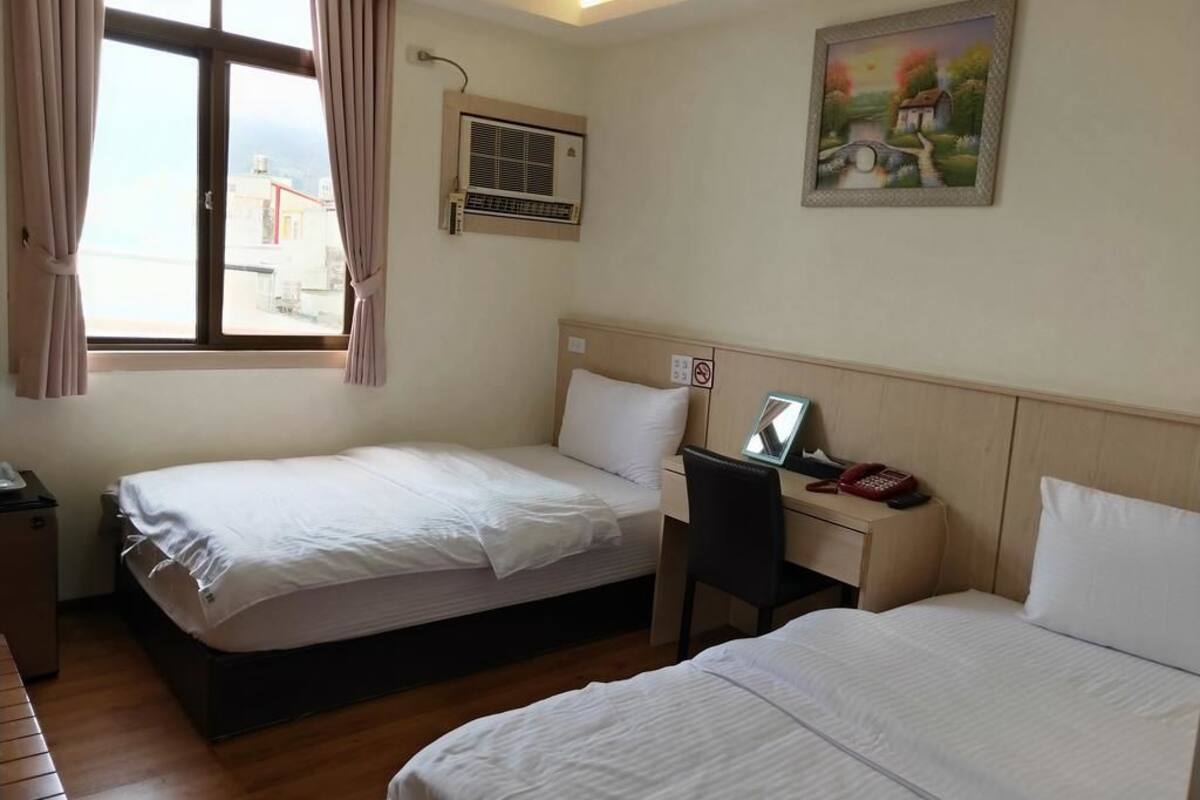 Habitación de hotel con dos camas individuales, un escritorio con silla, teléfono y ventana con cortinas.