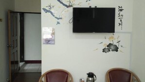 Una habitación con dos sillas de madera, una mesa con una tetera y tazas, un televisor en la pared y un mural decorativo.