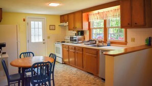 A compact kitchen with wooden cabinets, a microwave, a stove, and a sink. There is a dining table with chairs and a window with curtains. - Cozy 2 bedroom cottage less than a mile to the beach (Dennis Port)