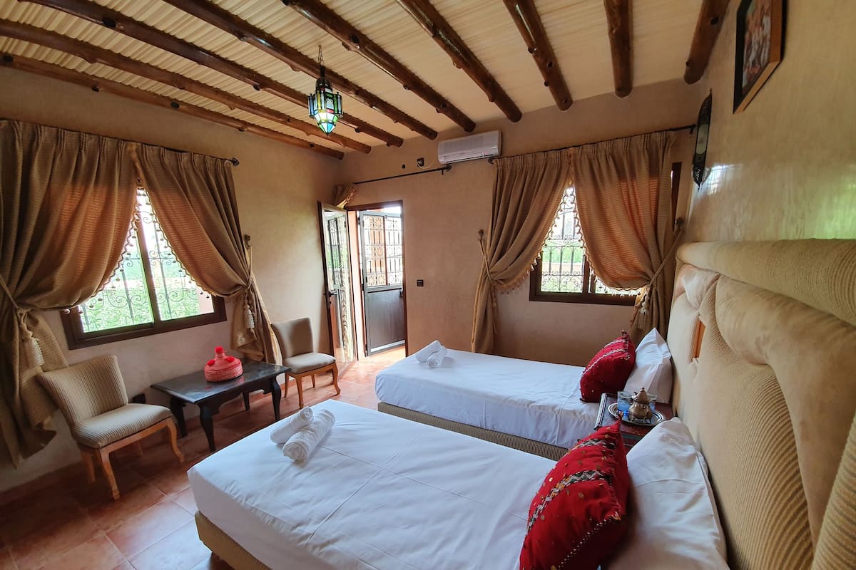 Quarto com duas camas, uma cadeira, uma mesa e uma janela com cortinas.